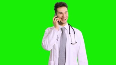 Yakışıklı doktor yeşil ekran krom anahtar arka planda izole edilmiş akıllı telefondan konuşuyor, pozitif gülümsüyor.