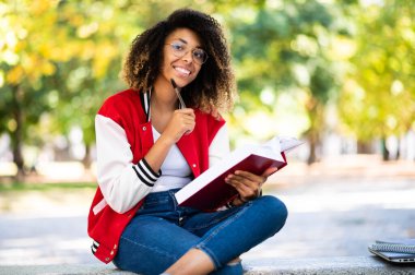 Güzel Afrikalı üniversite öğrencisi parkta bir bankta kitap okuyor.