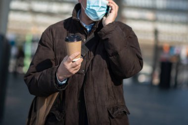 Covid Coronavirus konsepti, maskeli yaşlı iş adamı açık havada akıllı telefonunu kullanıyor.