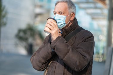 Covid Coronavirus konsepti, maskeli yaşlı iş adamı açık havada kahve içiyor.