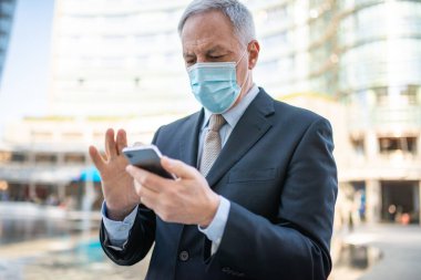 Covid konsepti, maskeli yaşlı iş adamı akıllı telefonunu açık havada bir şehirde kullanıyor.