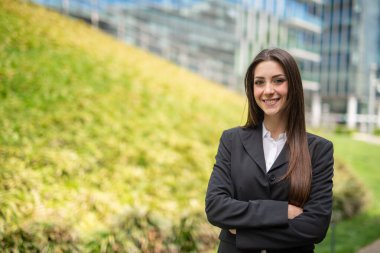 Çim tarlasının yanında gülümseyen genç bir kadın ve modern bir şehirde cam ofis binası.