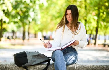 Güzel bir üniversite öğrencisi parkta bir bankta kitap okuyor.