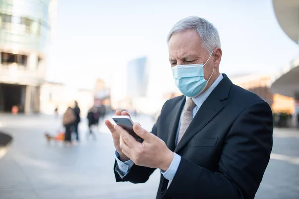 Covid coronavirus concept, masked elder businessman using his smartphone outdoor in a city