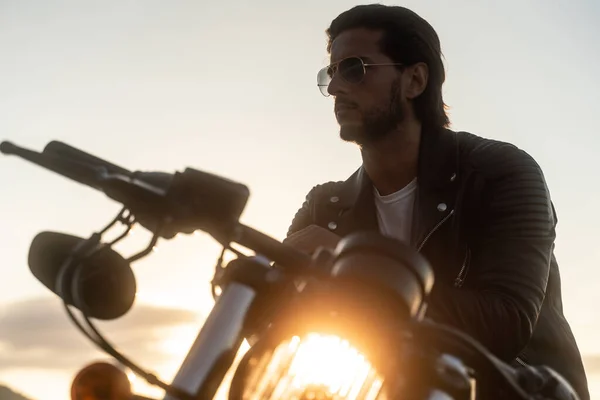 Motociclista guapo sentado en su motocicleta vintage, con chaqueta de cuero y gafas de sol Imagen de archivo