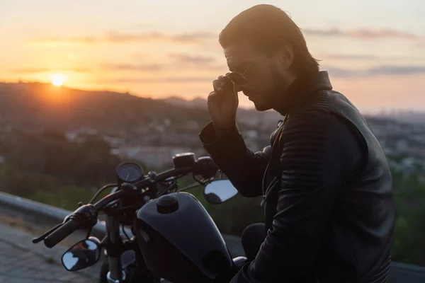 Retrato de verano del motorista sentado en su moto vintage, poniéndose gafas de sol al amanecer Fotos de stock