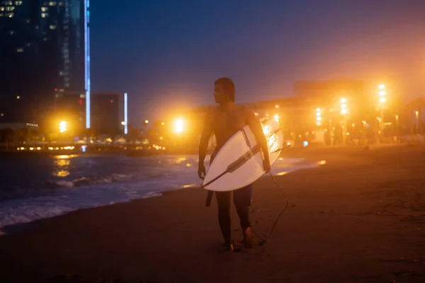 Foto nocturna de surfista joven sosteniendo tabla de surf y caminando por la costa, listo para el surf Imagen de stock