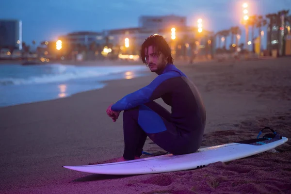 Retrato nocturno de surfista en traje de neopreno sentado en tabla de surf y mirando la cámara después de surfear Fotos de stock libres de derechos