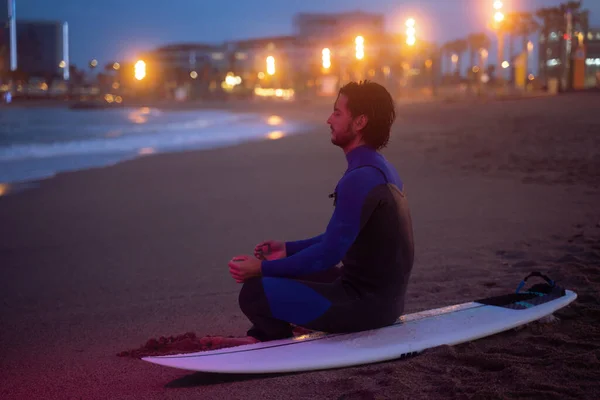 Retrato nocturno de surfista sentado en su tabla de surf y meditando en pose de loto después de surfear Fotos de stock libres de derechos