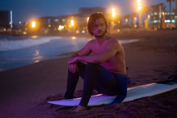Retrato nocturno del joven surfista sentado en su tabla de surf y mirando hacia otro lado después de surfear Imagen de archivo