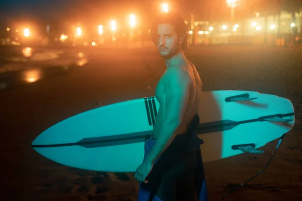 Retrato nocturno de surfista sosteniendo tabla de surf, de pie en luz verde neón, listo para el surf Fotos de stock libres de derechos