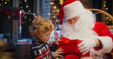 Noel Baba ve küçük beyaz çocuk Noel ağacının yanında dikilmiş, sihirli Noel hediyesini bekliyor. Kışın sıcak oturma odasında.