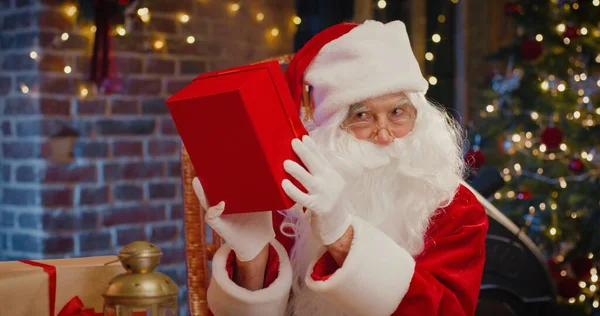 O que está dentro. Papai Noel barbudo tremendo com grande caixa de presente vermelho e adivinhe o que dentro enquanto passa o tempo no quarto com decorações de Natal no fundo. Conceito de espírito feriados — Fotografia de Stock