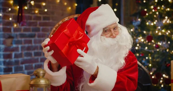O que está dentro. Papai Noel barbudo tremendo com grande caixa de presente vermelho e adivinhe o que dentro enquanto passa o tempo no quarto com decorações de Natal no fundo. Conceito de espírito feriados — Fotografia de Stock