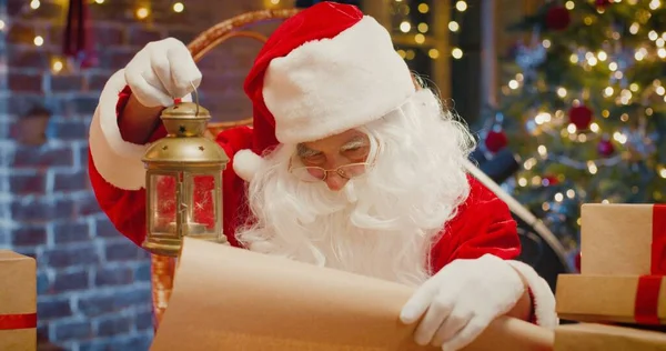 Papai Noel sentado na poltrona e lendo algo no papel enquanto segurava lanterna com decorações de Ano Novo no fundo. Conceito de espírito feriados — Fotografia de Stock