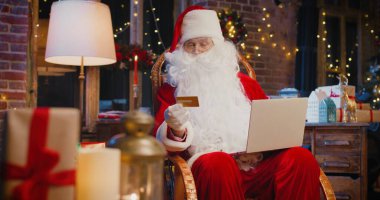 Neşeli, beyaz saçlı Noel Baba koltuğunda oturuyor ve laptopunda kredi kartıyla alışveriş yapıyor. Etrafta bir sürü Noel hediyesi ve hediye kutusu var. Tatil ruhu kavramı