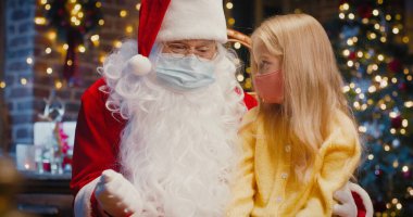 Noel Baba 'yla sırlarını paylaşırken dizlerinin dibinde oturan küçük sarışın kızın portresi. Koruyucu maskeler takıyorlar. Salgın konsepti