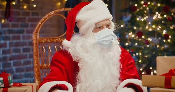 Télapó védőmaszkot visel ül a karosszékben, és mond valamit a kamerába, miközben otthon ül. Közelkép egy boldog végzős emberről Santa Clause jelmezben. Karácsonyi szellem koncepció — Stock Fotó