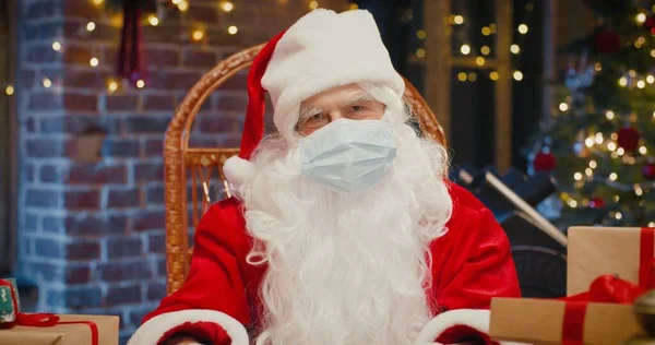 Санта-Клаус в защитной маске сидит в кресле и говорит что-то в камеру, сидя дома. Крупный план счастливого старшеклассника в костюме Санта Клауса. Концепция Рождества — стоковое фото