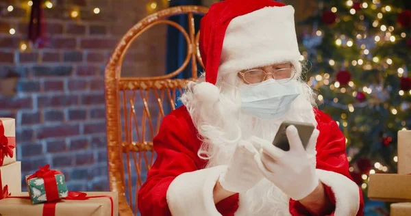 Papai Noel sentado na poltrona e usando seu smartphone enquanto sentado em casa com decorações de Ano Novo. Conceito de Natal e tecnologias — Fotografia de Stock