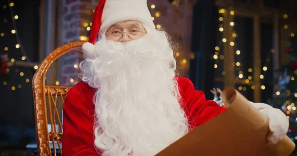 Papai Noel barbudo sentado na poltrona e examinando papel com presentes com decorações de Natal no fundo. Conceito de espírito feriados — Fotografia de Stock