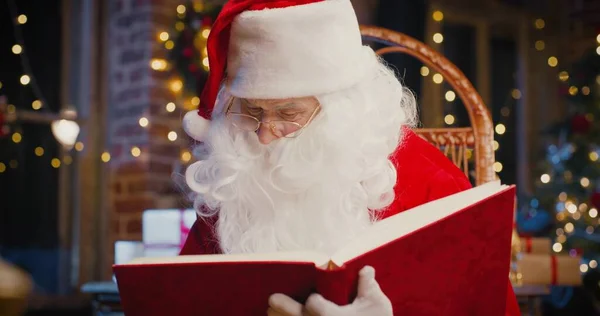 Papai Noel ler livro enquanto sentado na poltrona com presentes com decorações de Ano Novo no fundo. Conceito de espírito feriados — Fotografia de Stock