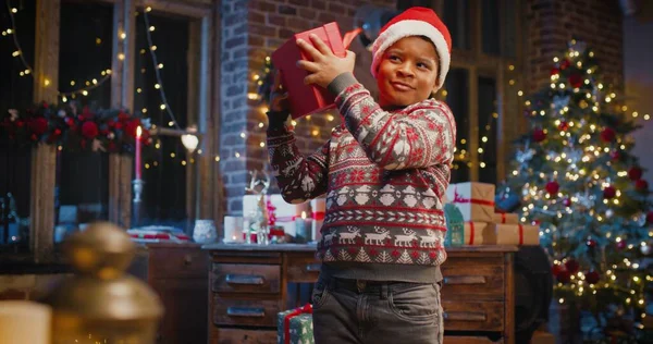 サンタの帽子の小さな多人種の子供の男の子ギフトボックスを保持し、バックグラウンドでクリスマスツリーとの贈り物を待っています — ストック写真