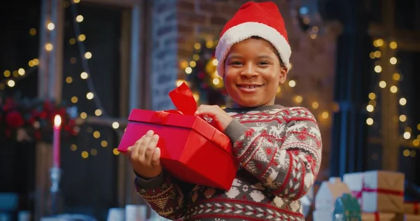 Kleine multiraciale jongen het dragen van Santa hoed houden geschenkdoos met cadeau, terwijl het doorbrengen van tijd thuis met Nieuwjaar decoraties. Kerstgedachte — Stockfoto