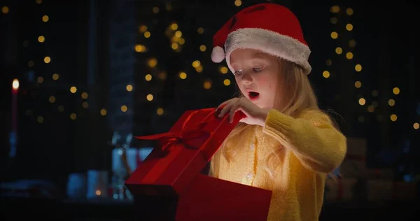 Kislány a Mikulás kalap nyitó ajándék doboz és kíváncsi az ajándék karácsonyfa a háttérben — Stock Fotó
