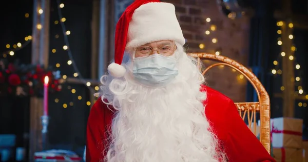 Homem alegre Papai Noel vestindo máscara protetora olhando para a câmera enquanto sentado em casa. Fechem-se os homens idosos felizes com o fato da Santa Cláusula. Conceito de espírito de Natal — Fotografia de Stock