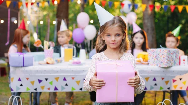 背後のテーブルに彼女の友人の多くと公園でカメラにポーズギフトボックスを持つ小さな白人ブロンドの女の子。誕生日おめでとう — ストック写真