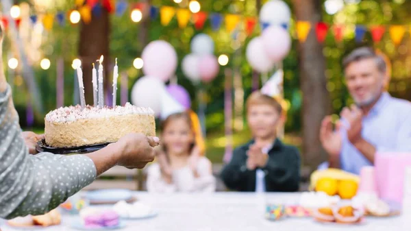 A mãe extrovertida que dá o bolo com velas ao filho alegre. Pai radiante sentado perto dele. A irmãzinha está a rir alto. Conceito de feliz aniversário — Fotografia de Stock