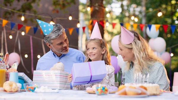 Es tan lindo. Pequeña cumpleañera sentada en la mesa y tomando juguete de oso de peluche de la caja de regalo mientras celebra en el jardín con sus padres — Foto de Stock