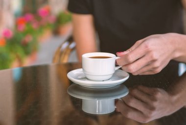 Bir kız kafede kahve içiyor. Kahve dükkanının arka planında, ahşap sehpanın üzerinde beyaz kahve fincanı.