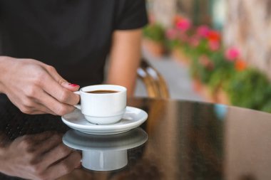 Bir kız kafede kahve içiyor. Kahve dükkanının arka planında, ahşap sehpanın üzerinde beyaz kahve fincanı.