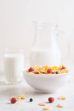 Bir kase mısır gevreği, sütlü, taze ahududu, yabanmersini. Sağlıklı kahvaltı için bir sürahi süt ve beyaz tahta üzerinde bir bardak.