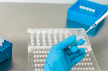 Bir laboratuvar teknisyeninin ellerinde bir tüp kan örneği ve diğer numunelerle dolu bir raf var. Üzerinde çalışmak için kan tüpü örneği tutan laboratuvar teknisyeni. Corona virüsü salgını konsepti. Coronavirus aşısı gelişimi.