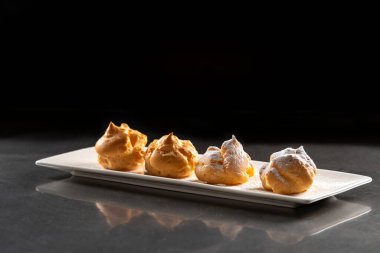 Profiterolleri pudra şekeriyle süslemek. Siyah arkaplanda krema şekerli ekler