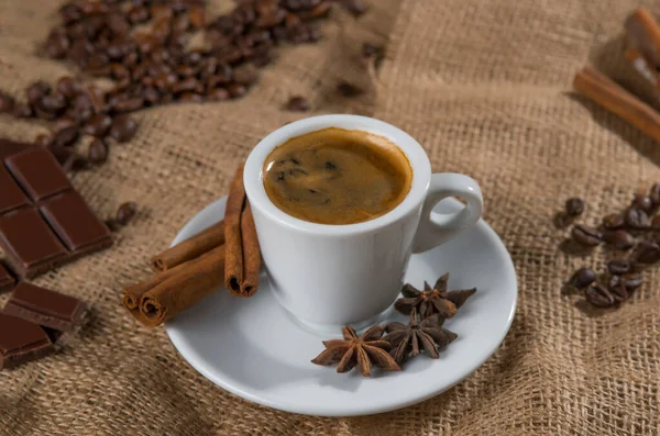 dark way of white cup of coffee with milk and aromatic spices: cinnamon sticks, star anise, dried slices of lemon and coffee beans on a napkin of burlap