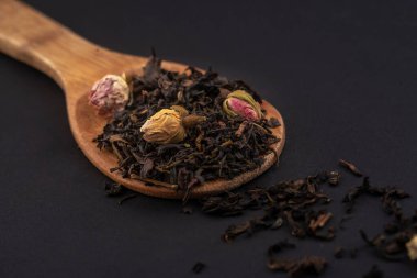 siyah arka plan üzerinde izole ahşap bir kaşık üzerinde gül çiçekleri ile aromatik siyah kuru çay