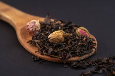 siyah arka plan üzerinde izole ahşap bir kaşık üzerinde gül çiçekleri ile aromatik siyah kuru çay