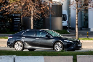 Moskova; Rusya - 14 Ekim 2021: Siyah Toyota Camry sıcak bir sonbahar gününde bir parkın arka planına doğru hızla sürüyor