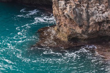 Dik, keskin bir kayaya yakın çekim. Denizin arka planında yeşillik var.