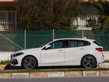 Yan, Türkiye 13 Şubat 2022: Beyaz BMW 1 serisi sıcak bir sonbahar gününde bir caddenin arka planına park edildi 