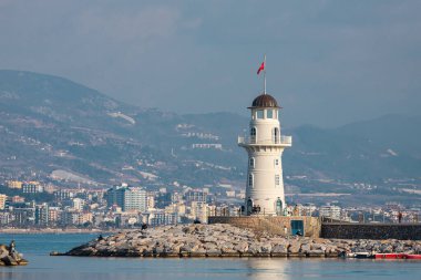 Alanya, Türkiye 16 Şubat 2022: Mavi denizin arka planında güzel beyaz bir deniz feneri ve birçok tekne ve yat bulunan bir rıhtım 