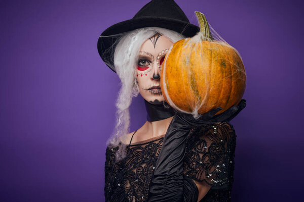 Серьёзная молодая женщина с хэллоуинским макияжем покрывает половину лица тыквой