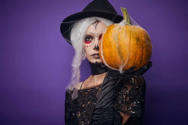 Mujer joven seria con maquillaje de Halloween cubre la mitad de la cara con calabaza Fotos de stock