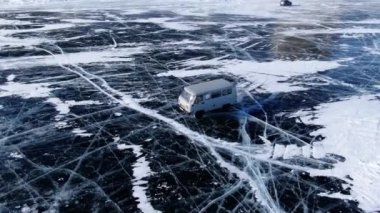 Sovyet beyaz arabası UAZ donmuş Baykal gölünün buzlu yüzeyinde yol alır. Yan görüş. Çatlaklı ve karlı mavi buz. Mükemmel panoramik görünüm.