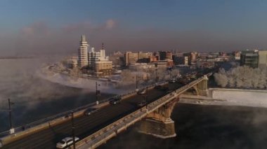 Angara nehrinin üzerindeki eski köprü. Buhar sudan yayılır. Hava aracı uçuşu. Kışın, Rus şehri buz ve karla kaplıdır..