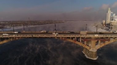 Angara nehrinin üzerindeki eski köprü. Buhar sudan yayılır. Hava aracı uçuşu. Kışın, Rus şehri buz ve karla kaplıdır..